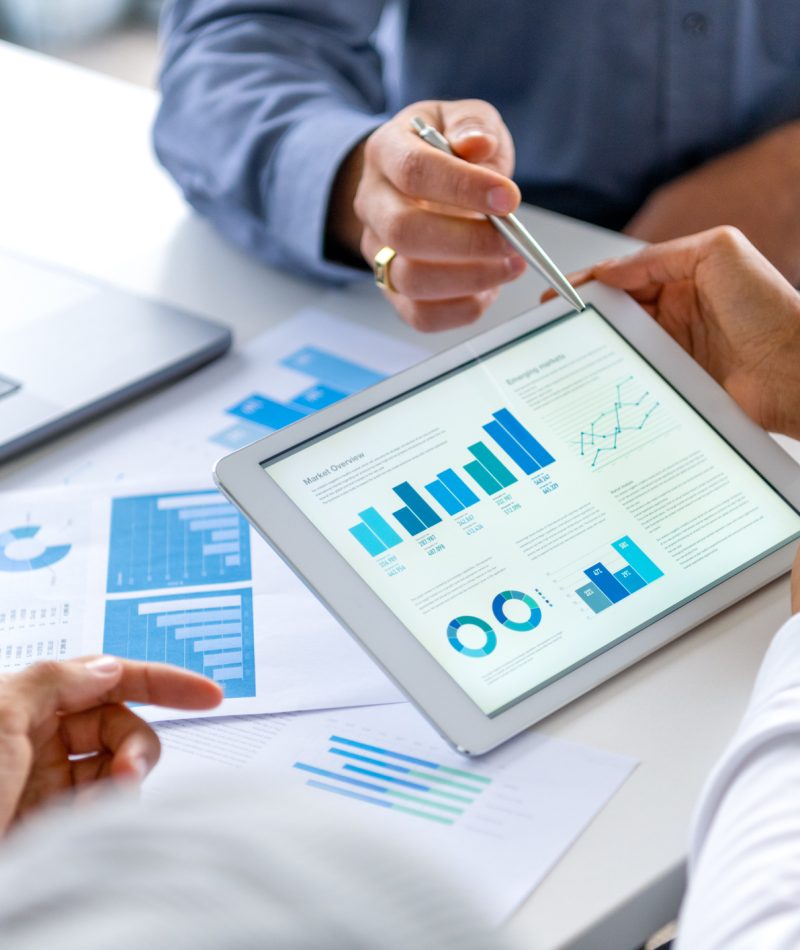 Close up of three people looking at financial data with graphs and charts.  All their hands can be seen and one person is pointing with a pen. There is paperwork on the desk showing more finance information
